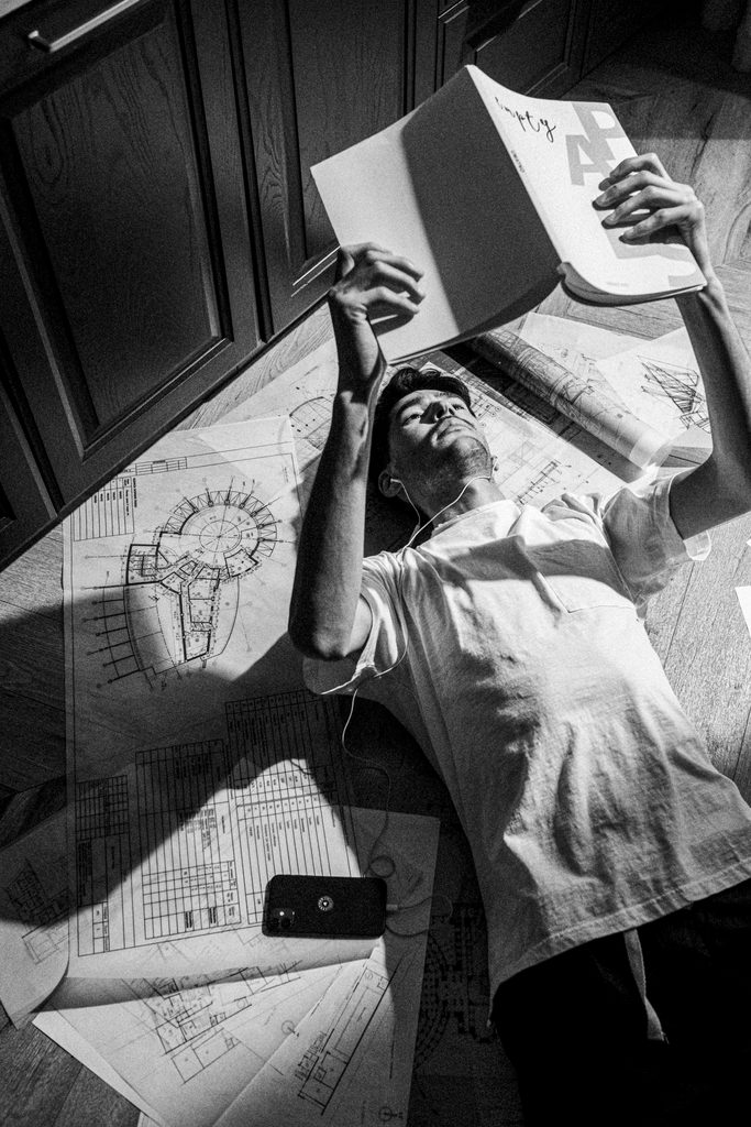 Young man lying among architectural drawings while looking through an open notebook and listening through earphones.